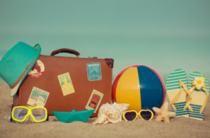 summer and vacations represented by a picture of a suitcase, beachball, sunglasses, flip-flops, and as seashell laying on beach sand